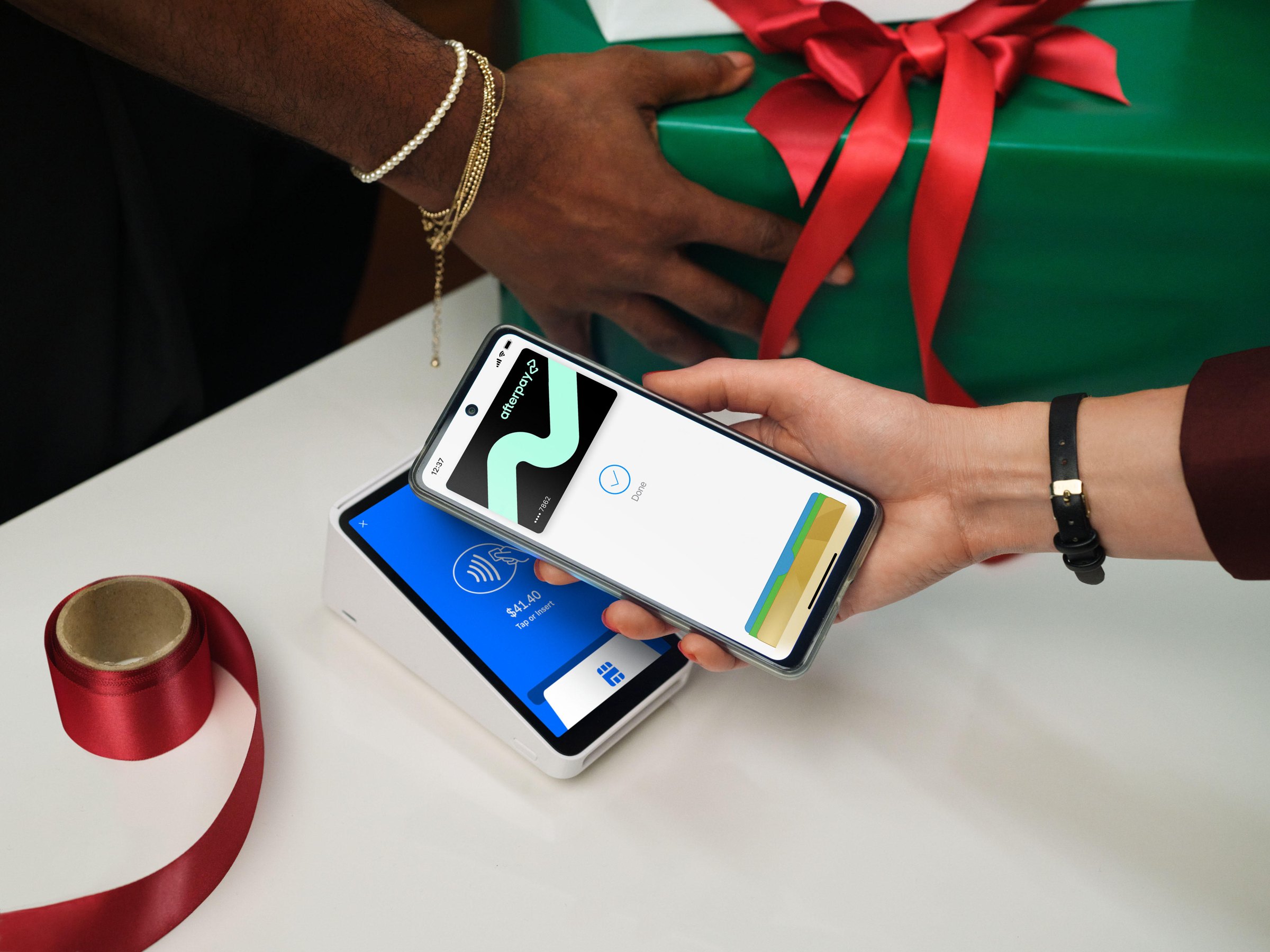 Hands holding a phone to a card terminal beside a green wrapped gift with a red ribbon