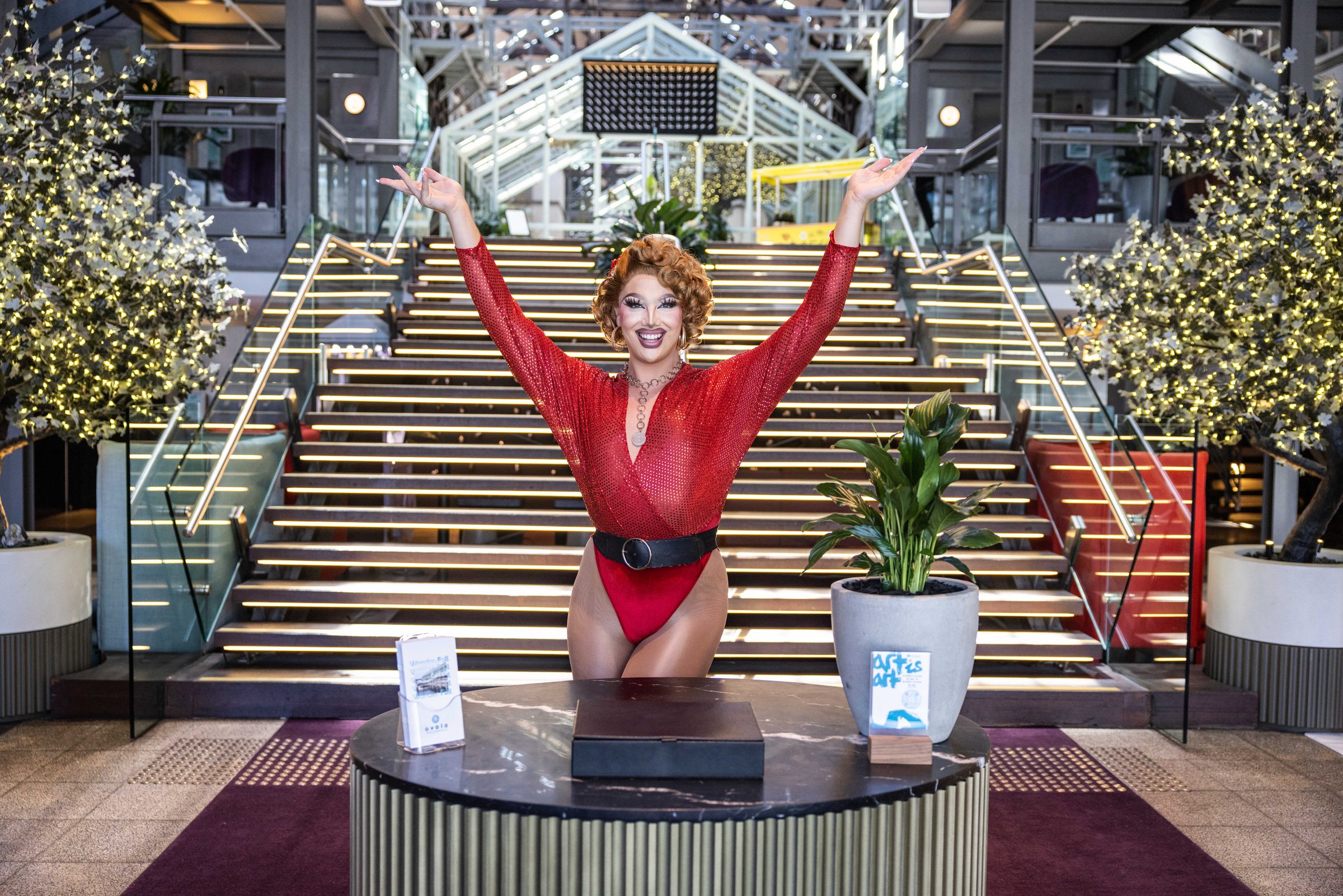 Performer in a red bodysuit, arms raised, on a plinth in front of an Ovolo hotel staircase